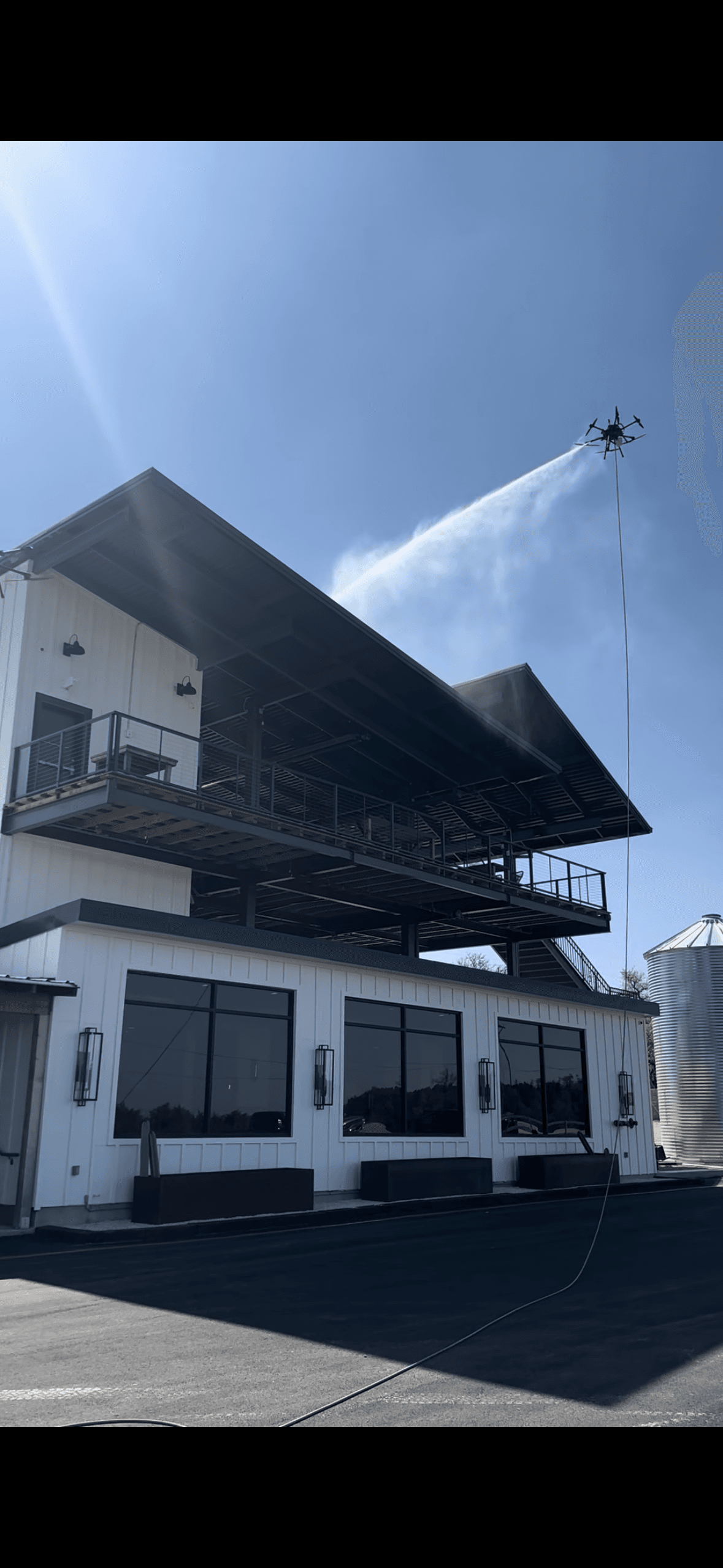 AES drone hovering above a 2-story building, actively cleaning the exterior on a sunny day